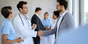 Male doctor handshaking with a businessman while meeting in a hallway at medical clinic.